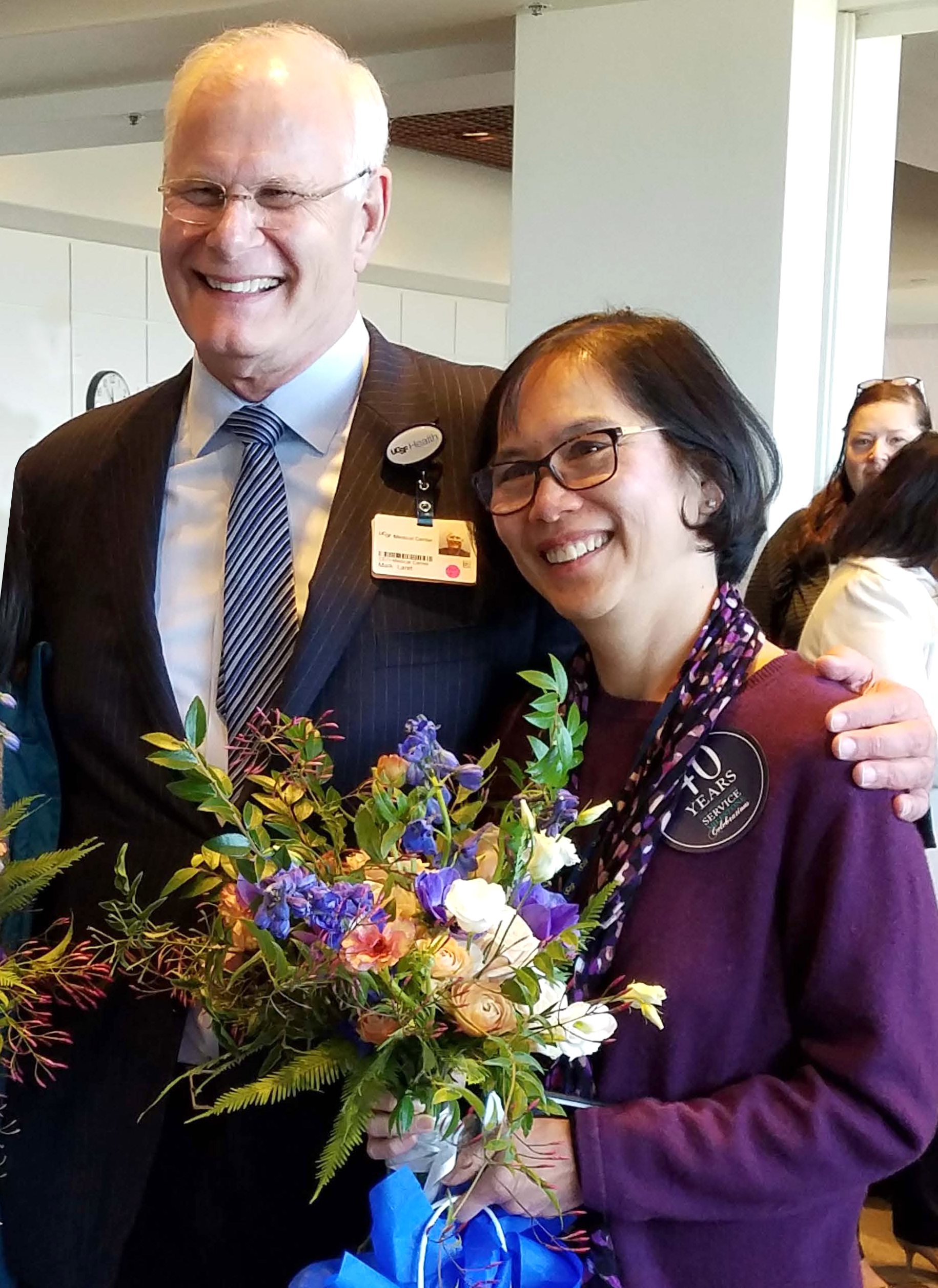 Two smiling people standing together indoors at a recognition event. One person holds a colorful bouquet of flowers while the other has an arm around them. Both are wearing badges, and one badge notes years of service. The background shows other attendees and a bright, open interior space.
