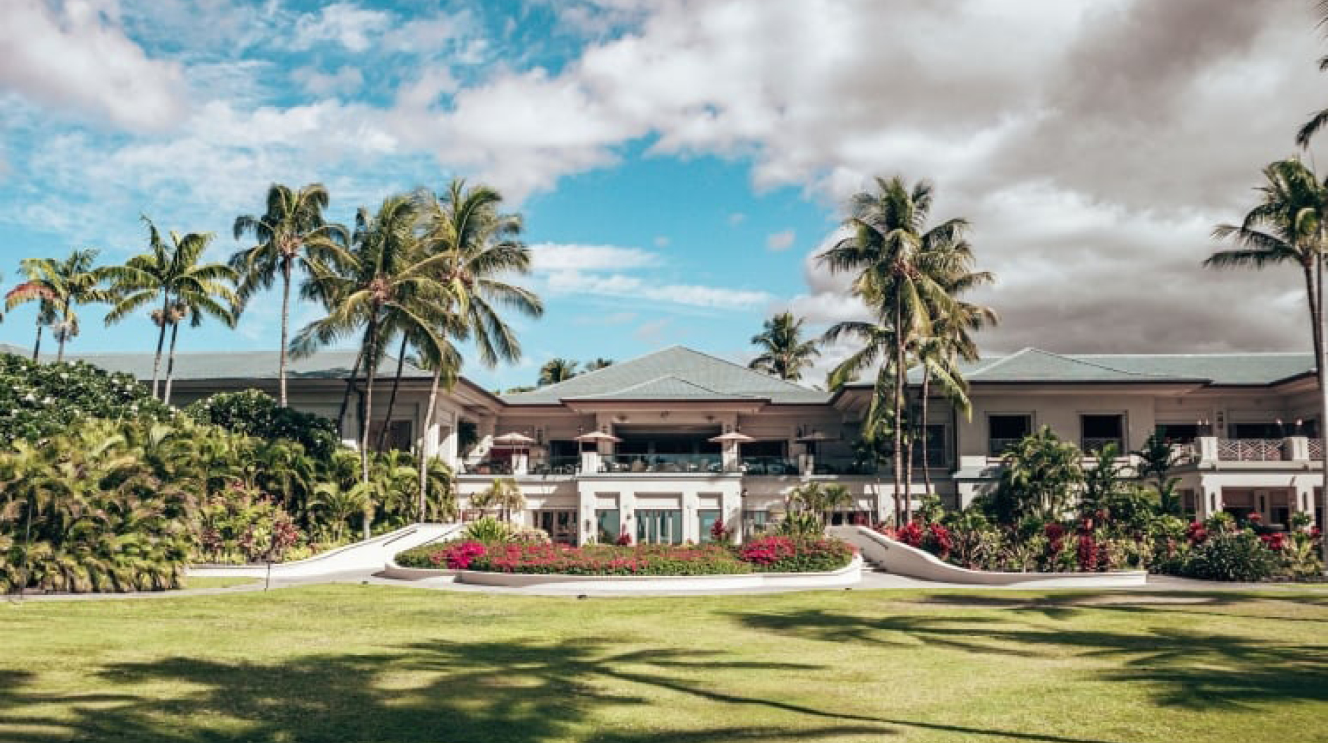 Exterior view of the Fairmont Orchid Resort & Spa featuring a low, elegant resort building framed by tall palm trees, lush tropical landscaping, and a wide green lawn under a partly cloudy blue sky.