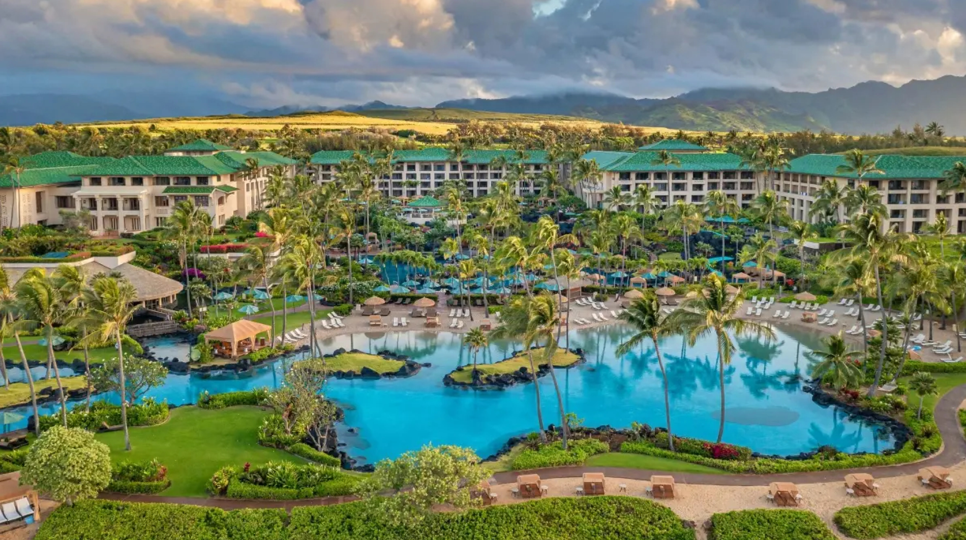 Aerial view of the Grand Hyatt Kauai Resort & Spa featuring expansive lagoon-style pools surrounded by palm trees, lounge chairs, and landscaped gardens, with resort buildings and distant mountains under a dramatic cloudy sky.