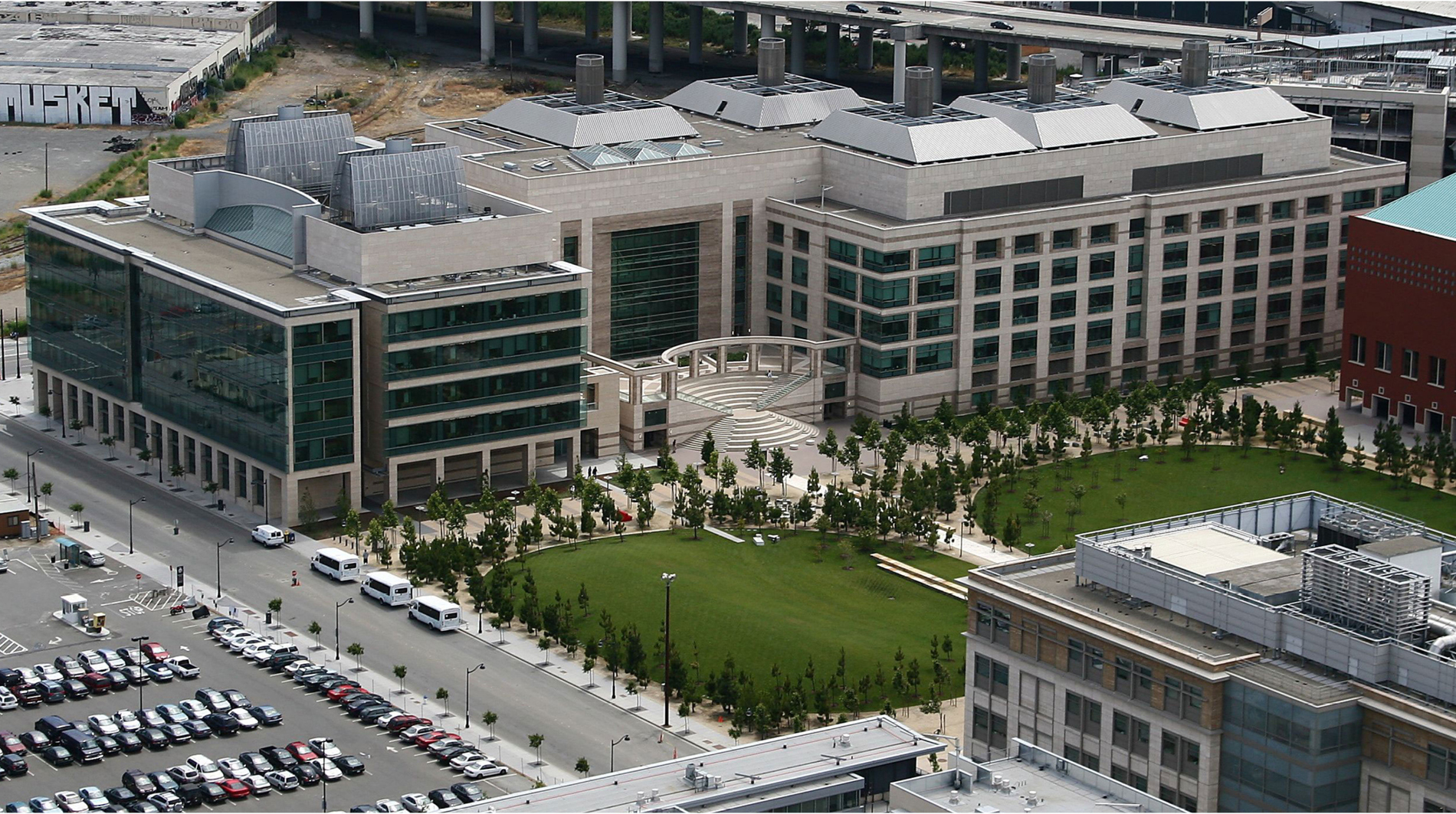 Aerial view of a large modern campus building with glass and stone facades, surrounded by landscaped green lawns, rows of trees, nearby streets, and parking areas in an urban setting.
