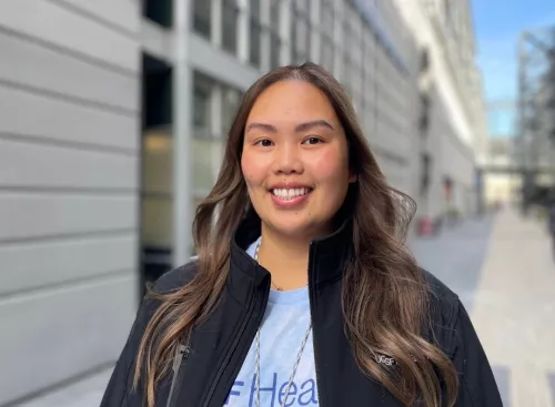 UCSF Health staff member smiling outdoors in an urban courtyard with modern buildings in the background. She is wearing a light blue UCSF Health shirt and a black jacket, with natural light highlighting her friendly expression.