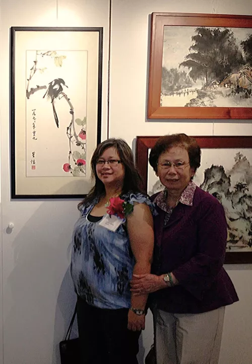 Two women stand closely together in front of framed artwork at an indoor gallery. The younger woman on the left wears glasses and a blue patterned top with a red corsage. The older woman on the right rests her hand on the younger woman’s arm and wears a purple jacket and glasses. Paintings with nature scenes hang on the wall behind them.