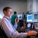 Radiologist reviewing medical imaging scans on multiple computer monitors in a dimly lit reading room. He is holding a microphone for dictation while analyzing a CT scan displayed on one of the screens, with another medical professional working in the background.