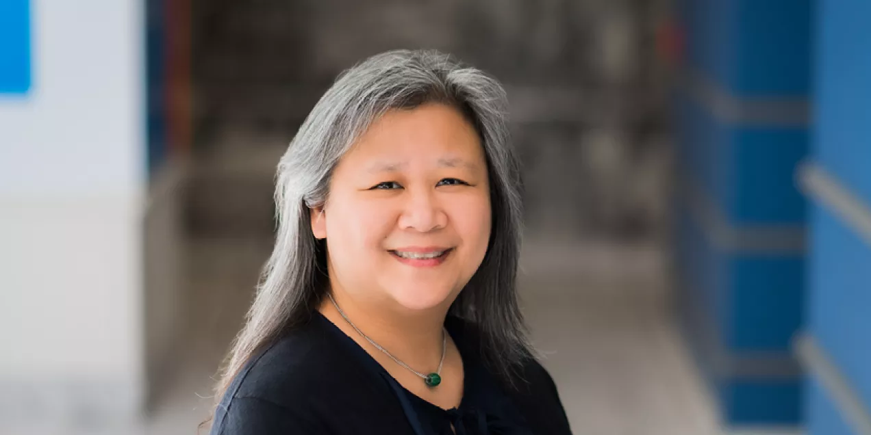 Cindy Cheng with long gray hair smiles softly while seated in a bright indoor hallway. She is wearing a dark top and a green pendant necklace, with blurred architectural details in the background.