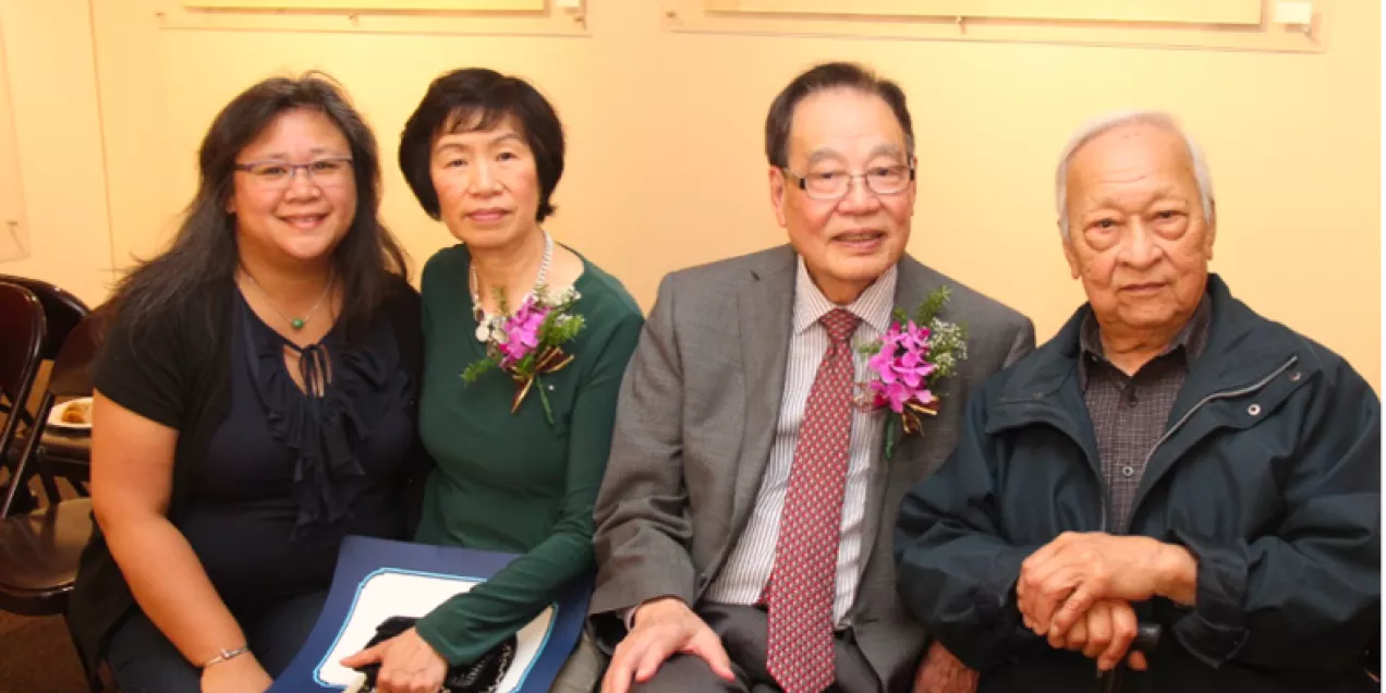 Four people sit closely together at an indoor event, smiling toward the camera. A woman in dark clothing sits on the left, next to a woman in a green top wearing a floral corsage. Beside them is an older man in a suit with a matching corsage, and another older man in a dark jacket holding a cane. Framed artwork hangs on the wall behind them.