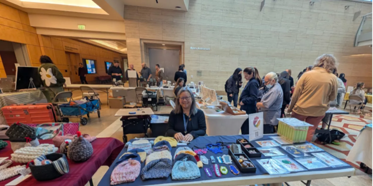 A woman sits behind a craft table at an indoor artisan fair, smiling at the camera. Her table displays handmade knitted hats, headbands, and small crafts. Other vendors and visitors fill the bright, spacious lobby around her, with natural light coming through large windows above.