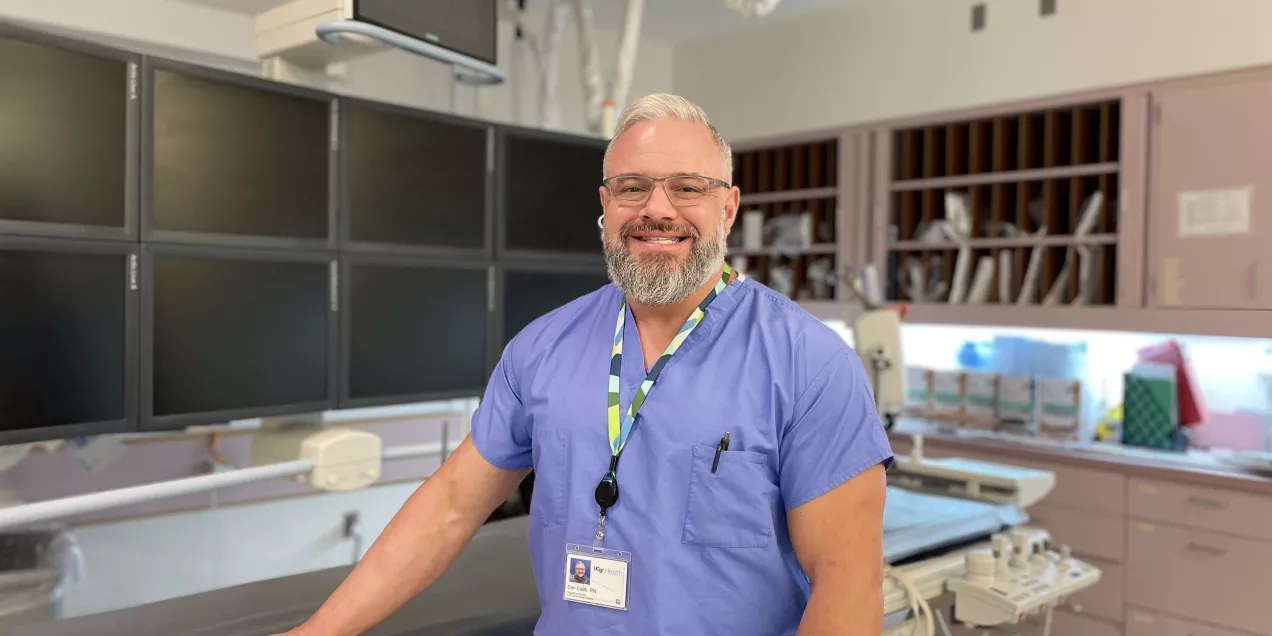A medical professional in blue scrubs standing in a clinical imaging room, smiling at the camera. He rests one hand on the exam table, with multiple blank monitors behind him and medical equipment and cabinetry in the background.