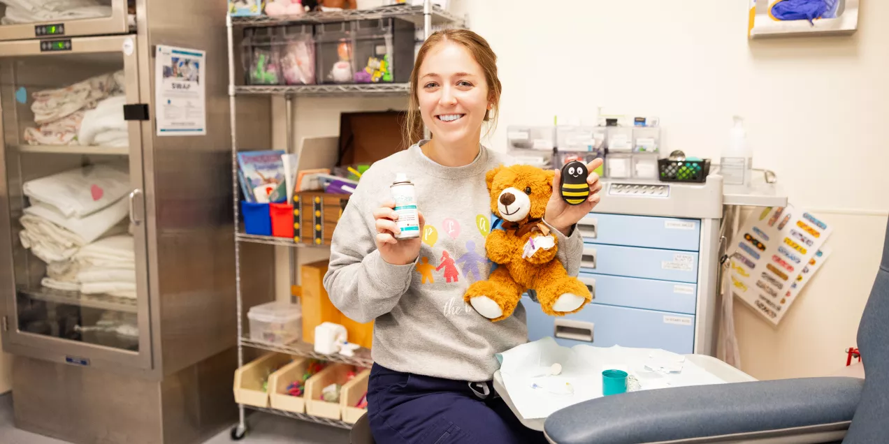 Emily Keis, a child life specialist sitting in a clinical room, smiling while holding a stuffed teddy bear in one hand and a small bee-shaped toy in the other. Medical supplies, toys, and storage cabinets are visible in the background.