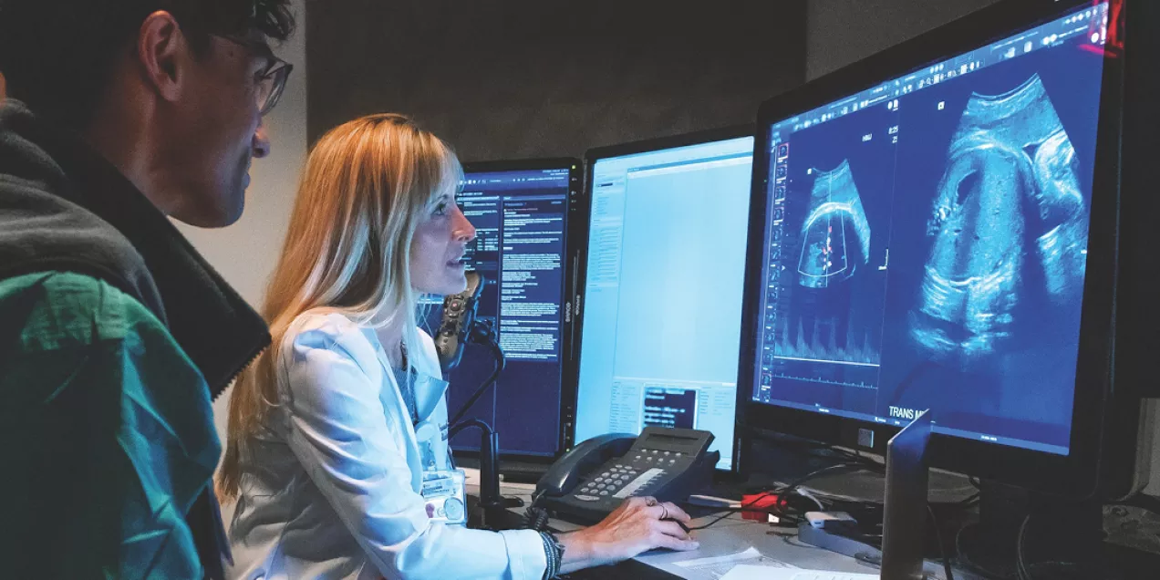 Lori Strachowski and a clinician at a workstation reviewing ultrasound images on multiple large monitors, while another person stands beside her observing. The room is dimly lit, and the screens display detailed medical imaging.