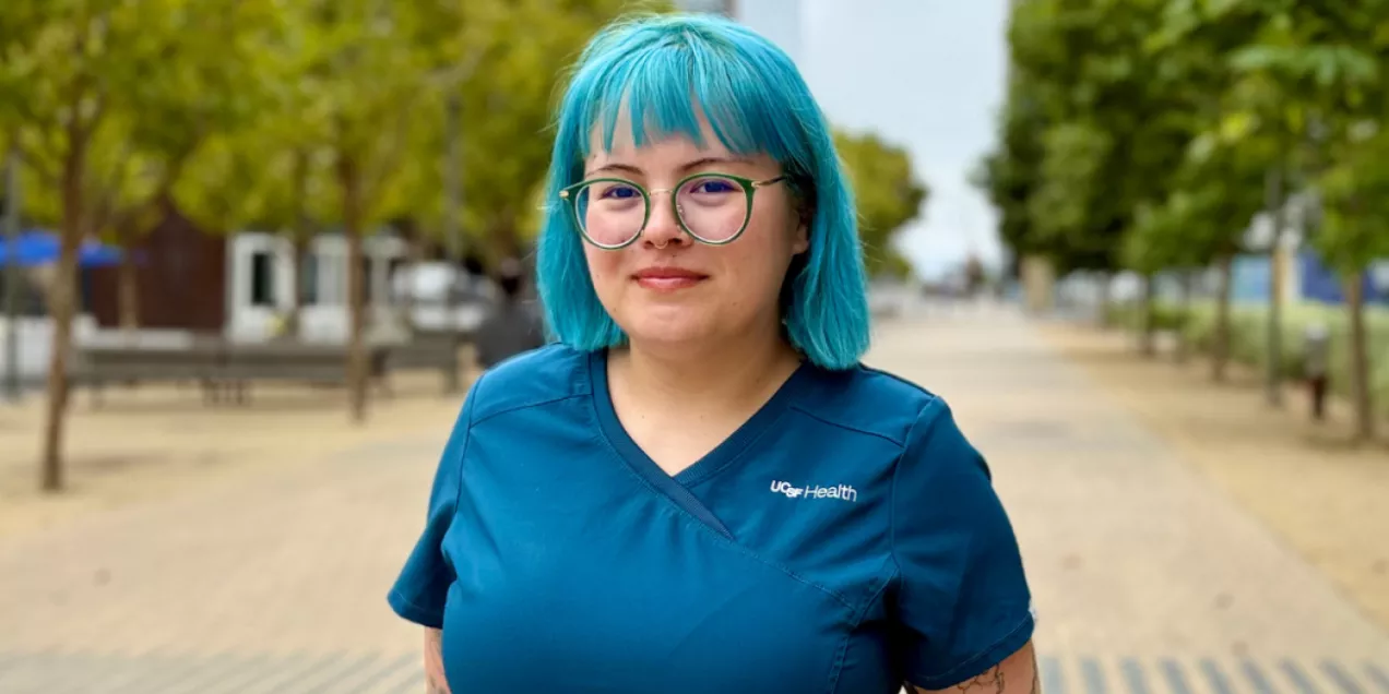 A person with teal hair and glasses stands outdoors on a pathway lined with trees and buildings in the background. They are wearing teal scrubs with a UCSF Health logo and have tattoos on both arms, smiling softly at the camera.