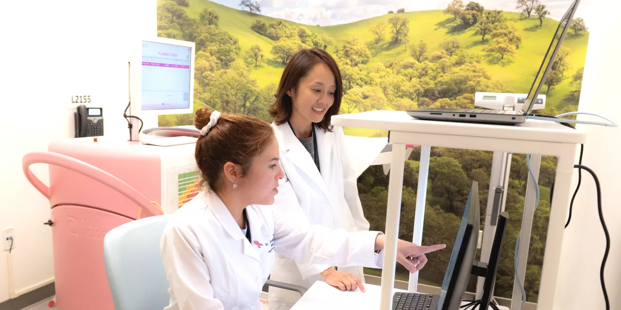 Two clinicians in white lab coats working at a computer workstation, with one seated and pointing at the monitor while the other stands beside her. Medical equipment and a wall mural of green hills are visible in the background.