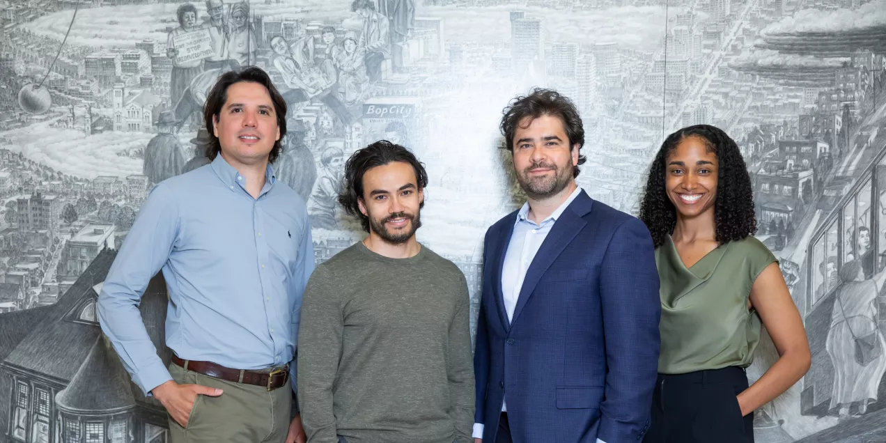Four people standing side by side and smiling in front of a large, detailed grayscale mural depicting a cityscape and historical scenes.