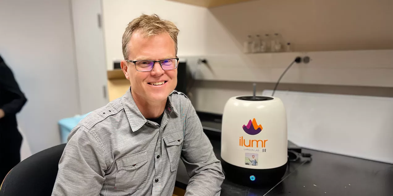 seated in a lab next to a small tabletop MRI device labeled ilumr, smiling toward the camera in a workspace with equipment in the background