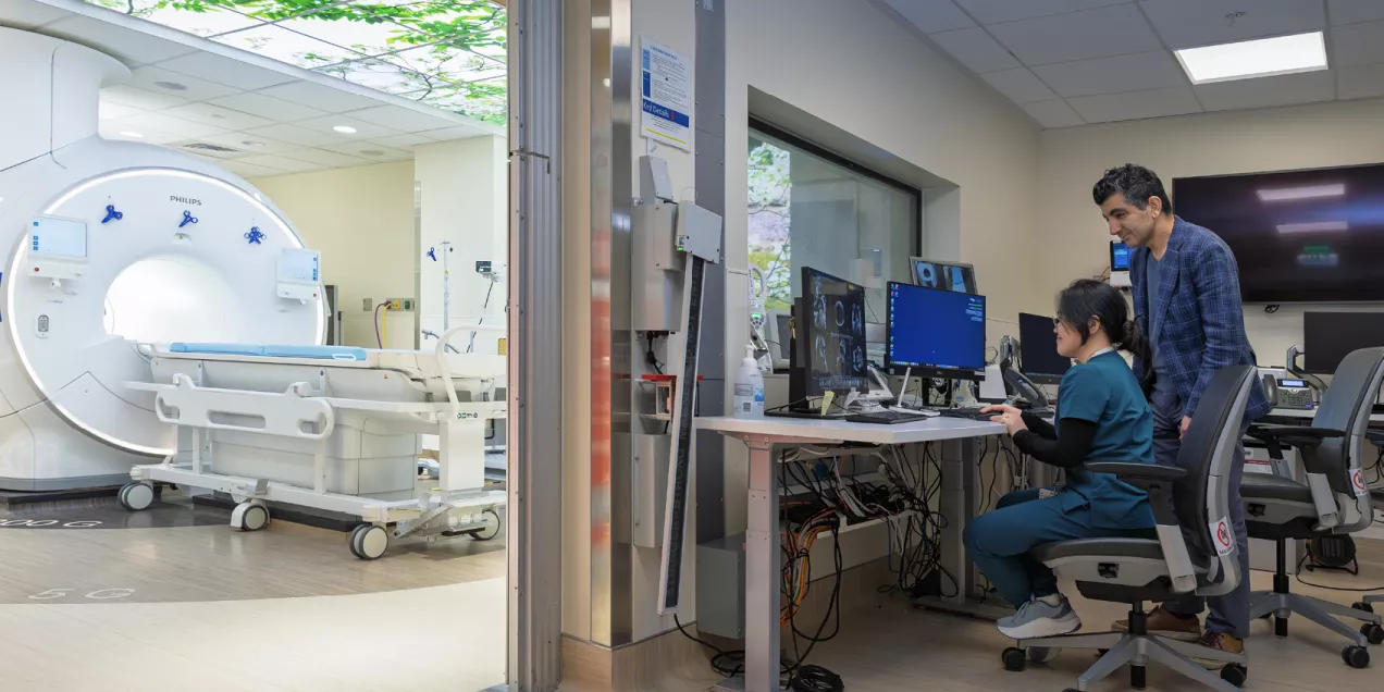 A large MRI scanner in a bright, modern imaging room is shown on the left, while two medical professionals work at computer monitors in an adjacent control room on the right. The scene includes medical equipment, monitors displaying scans, and a ceiling light panel with a leafy design.