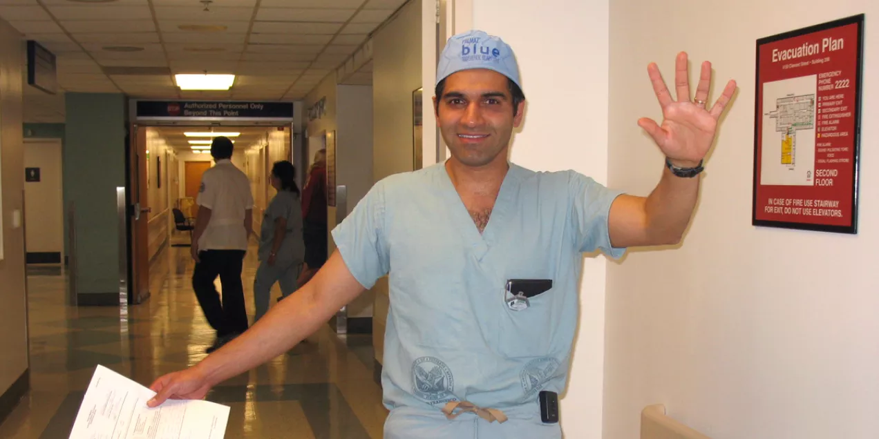 Rajiv Sawhney wearing light blue scrubs and a surgical cap stands in a hospital hallway, holding papers in one hand and waving with the other. Several people are visible in the background walking down the corridor.