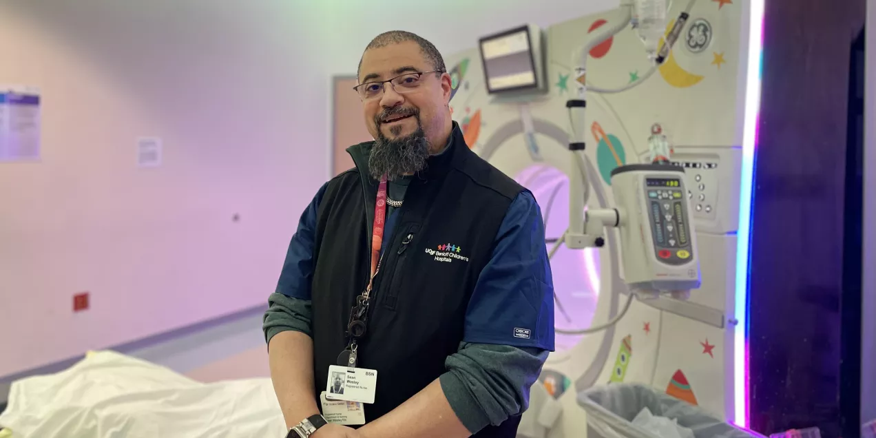 A smiling man with glasses and a goatee stands in a brightly lit pediatric hospital imaging room. He’s wearing a dark vest with the UCSF Benioff Children’s Hospitals logo and a colorful badge lanyard. Medical equipment and playful wall designs are visible in the background.