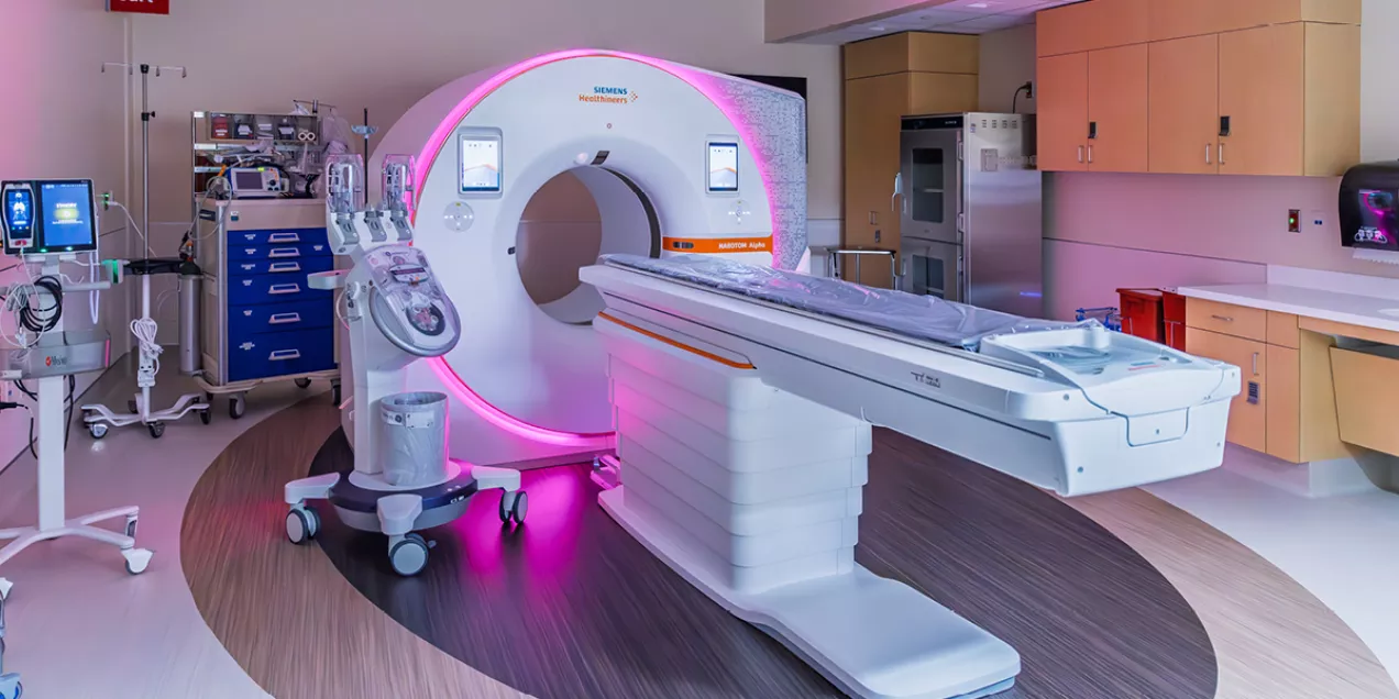 Photon-counting CT imaging suite with a Siemens scanner glowing with pink accent lighting, surrounded by medical equipment, cabinets, and a patient table in a clean clinical room