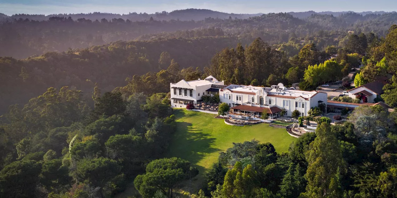 A large hilltop resort building surrounded by dense green forest, with terraced lawns in front and rolling wooded hills stretching into the distance under soft afternoon light.