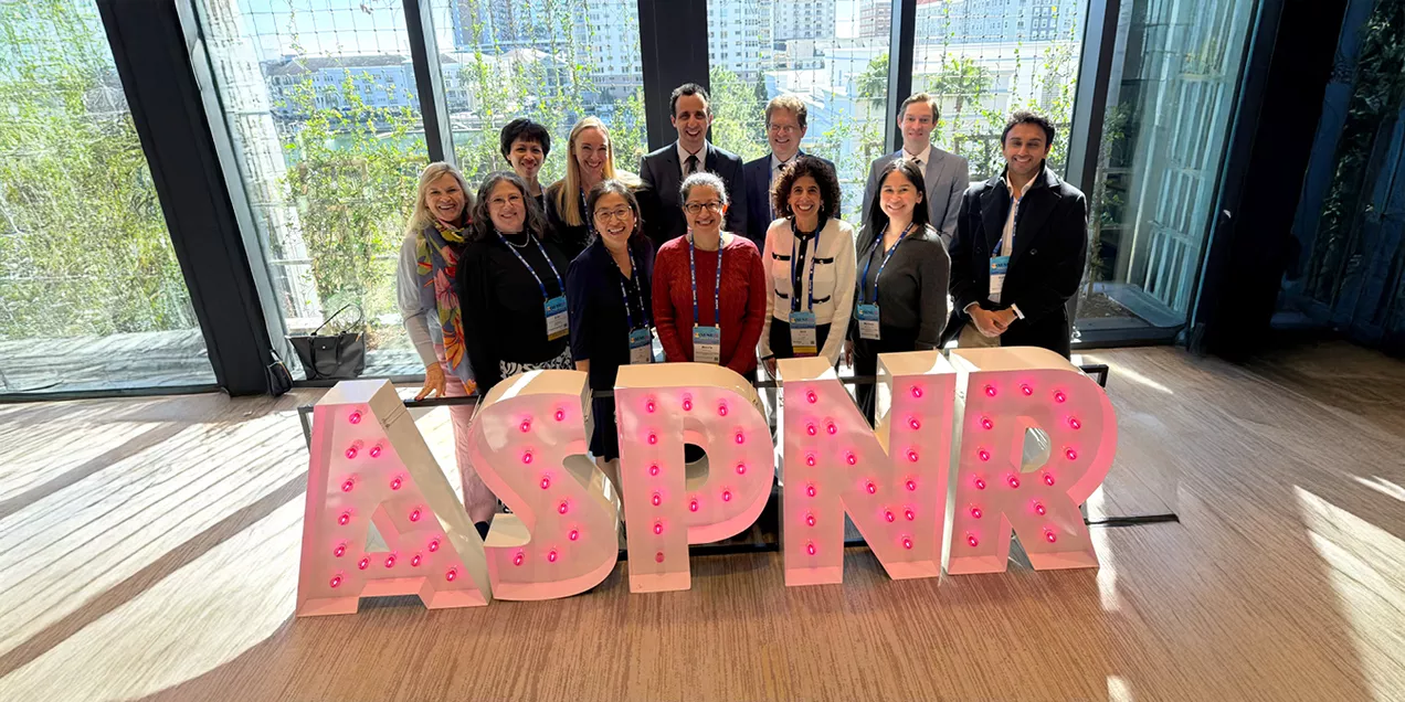 Group photo of UCSF faculty, alumni, and trainees standing indoors behind large illuminated ASPNR letters, smiling at the camera, with conference badges visible and a city view through floor to ceiling windows in the background.