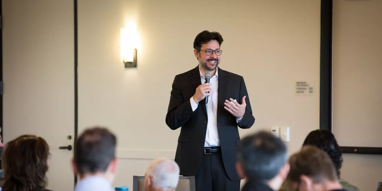 Christopher Hess dressed in a suit holds a microphone and addresses an audience during a professional presentation in a conference room.