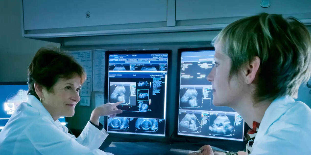 A clinician stands in a radiology reading room, smiling toward the camera, with imaging workstations visible behind them.