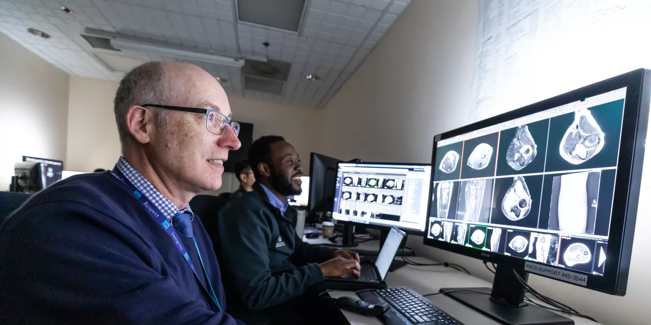 Two clinicians sit at workstations in a radiology reading room, reviewing medical imaging scans displayed on large computer monitors.