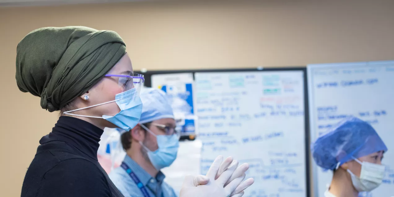 Clinicians wearing masks and gloves stand together in a clinical setting, preparing for a radiology procedure with whiteboards visible in the background.