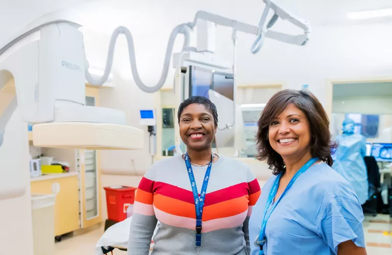 Two UCSF Health radiology staff members standing in a cardiovascular imaging procedure room beside a fluoroscopy imaging system, smiling and wearing ID badges and clinical attire.