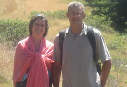 couple standing outdoors on a sunny trail, holding hands and smiling with trees and dry grass behind them
