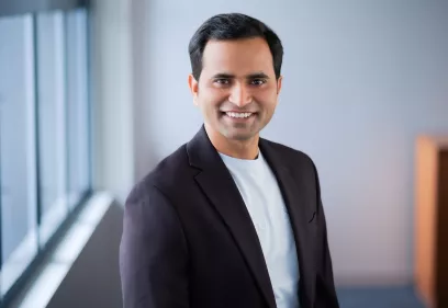 Anil Bidkar smiling, wearing a dark blazer and light shirt, standing near a window with a blurred background. The UCSF logo appears on the right against a dark blue backdrop.