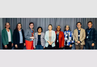 Group of nine professionals standing in a row in front of a gray curtain, smiling at the camera. They are dressed in business and professional attire, wearing a mix of jackets, blazers, and ID badges, representing a formal or academic gathering.