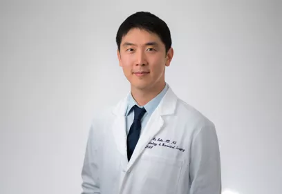Jae Ho Sohn Portrait wearing a white lab coat and navy tie, standing against a light gray background.