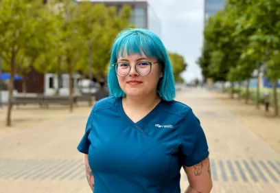 A person with teal hair and glasses stands outdoors on a pathway lined with trees and buildings in the background. They are wearing teal scrubs with a UCSF Health logo and have tattoos on both arms, smiling softly at the camera.