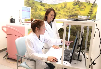 Two clinicians in white lab coats working at a computer workstation, with one seated and pointing at the monitor while the other stands beside her. Medical equipment and a wall mural of green hills are visible in the background.