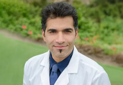 Sawhney Rajiv wearing a white lab coat, navy shirt, and blue tie, standing outdoors with a blurred background of greenery and flowers. He is looking at the camera with a slight smile.