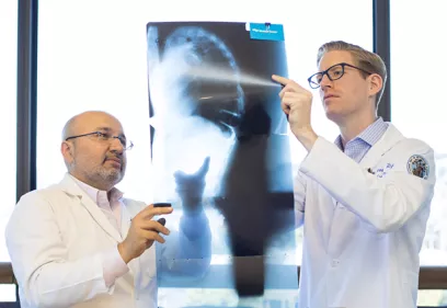 two doctors in white coats reviewing a large spinal X-ray held up to the light, discussing findings while standing in front of a bright window