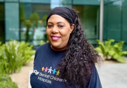 Ingrid Mcgraw smiling with long curly hair wearing a black headband and a navy UCSF Benioff Children’s Hospitals shirt stands outdoors in front of a modern glass building with greenery in the background.