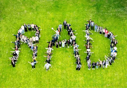 A large group of peopl e standing on a grassy field arranged to form the letters RAD, seen from above. The group is smiling and looking up with arms raised, creating a playful and energetic formation.