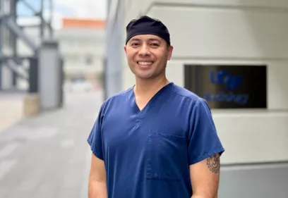 Portrait of a UCSF nurse wearing blue scrubs and a black surgical cap, smiling outdoors near a medical facility entrance.