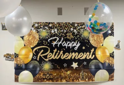 Decorative retirement banner reading “Happy Retirement” with gold and black balloons, confetti, stars, and celebratory accents displayed on a wall.