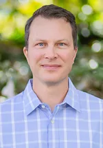 Theodore Jerdee outdoors wearing a light blue checkered button up shirt, looking at the camera with a neutral, approachable expression.
