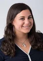 Samira Zebarjadian with long dark wavy hair, wearing a dark top and a necklace, smiling against a neutral background.