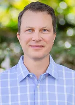 Theodore Jerdee outdoors wearing a light blue checkered button up shirt, looking at the camera with a neutral, approachable expression.