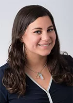 Samira Zebarjadian with long dark wavy hair, wearing a dark top and a necklace, smiling against a neutral background.