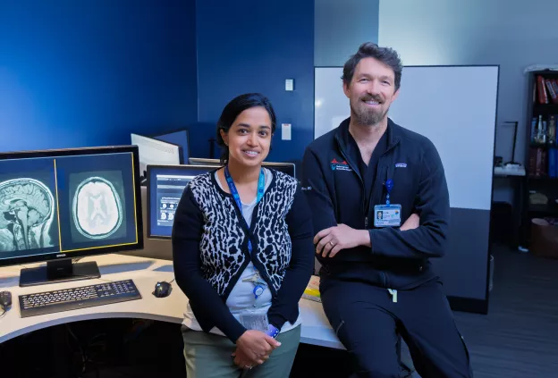 Two clinicians stand together in a radiology reading room beside computer monitors displaying brain imaging studies, smiling toward the camera.
