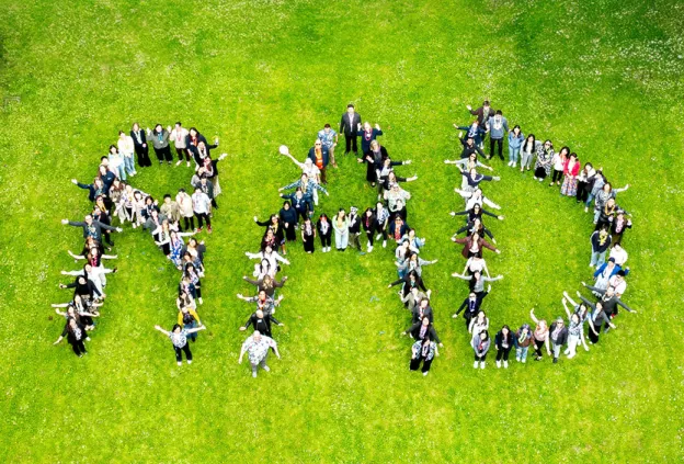 A large group of peopl e standing on a grassy field arranged to form the letters RAD, seen from above. The group is smiling and looking up with arms raised, creating a playful and energetic formation.