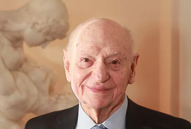 An elderly man in a suit and tie smiles toward the camera in a formal portrait setting.