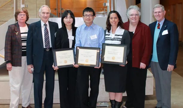 Group of eight people posing indoors. Three individuals in the center hold framed certificates, while others stand on either side of them smiling.