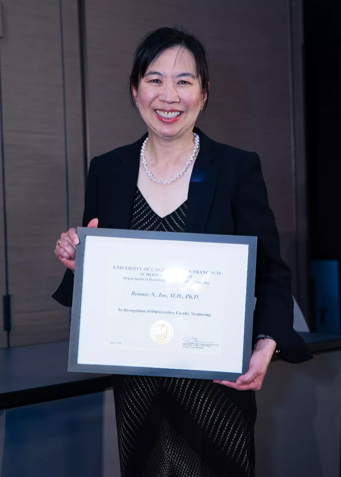 Person smiling indoors while holding a framed certificate toward the camera, wearing a black blazer and necklace.