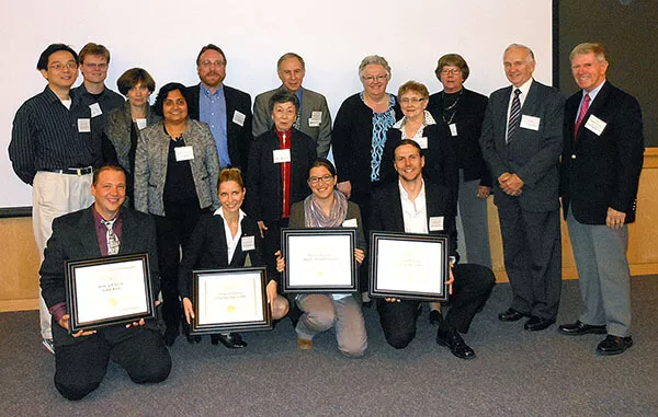 Large group of people posing indoors for a group photo. Four individuals in the front row are kneeling and holding framed certificates, while others stand behind them smiling.
